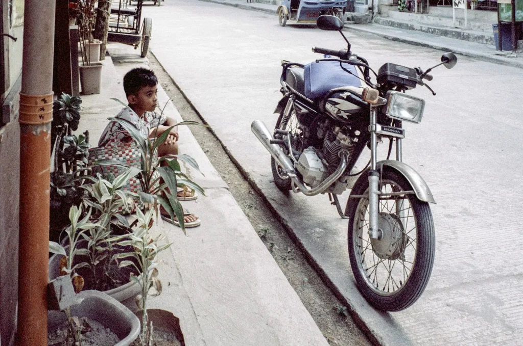 A young boy sits on the ground, his arms rested on his knees, as a blue motorcyle is parked in front of him.