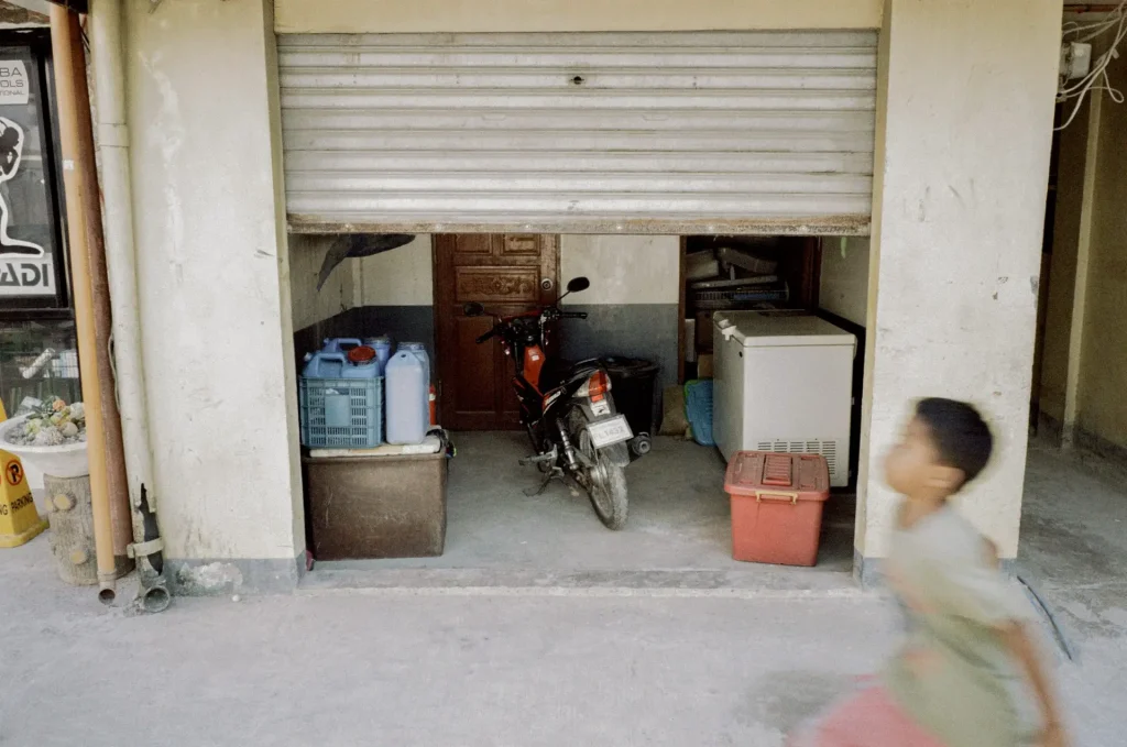 A young boy runs past a garage with its door half way open, revealing a parked motorcycle.