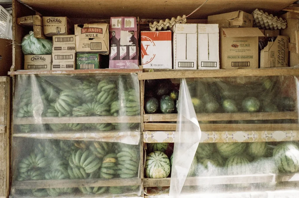 Watermelon and other wares are lined up on wooden shelves in an outdoor vendor stall.