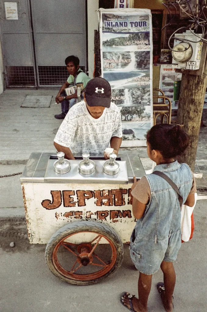 A vendor sells ice cream to a young girl as a man sits behind them looking at the photographer.