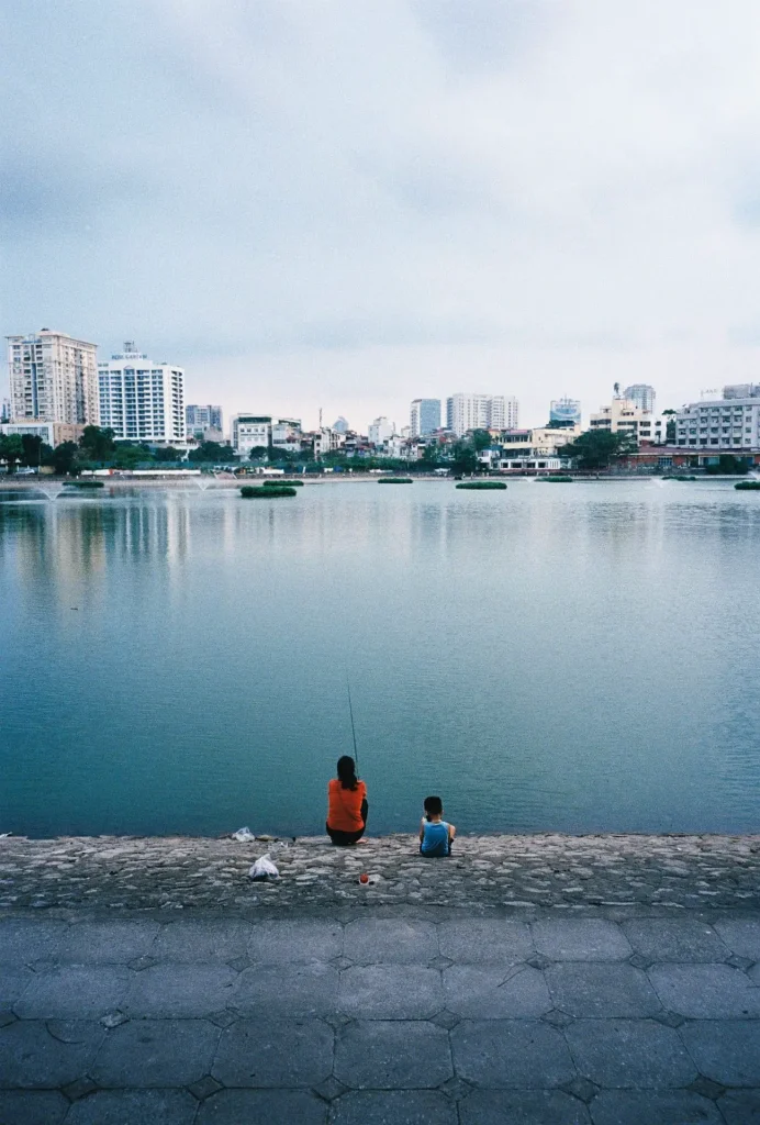 A woman in a red sweater fishes in Giang Vo lake next to her sun in a blue shirt. Buildings reflect of the lakes aquamarine surface. 
