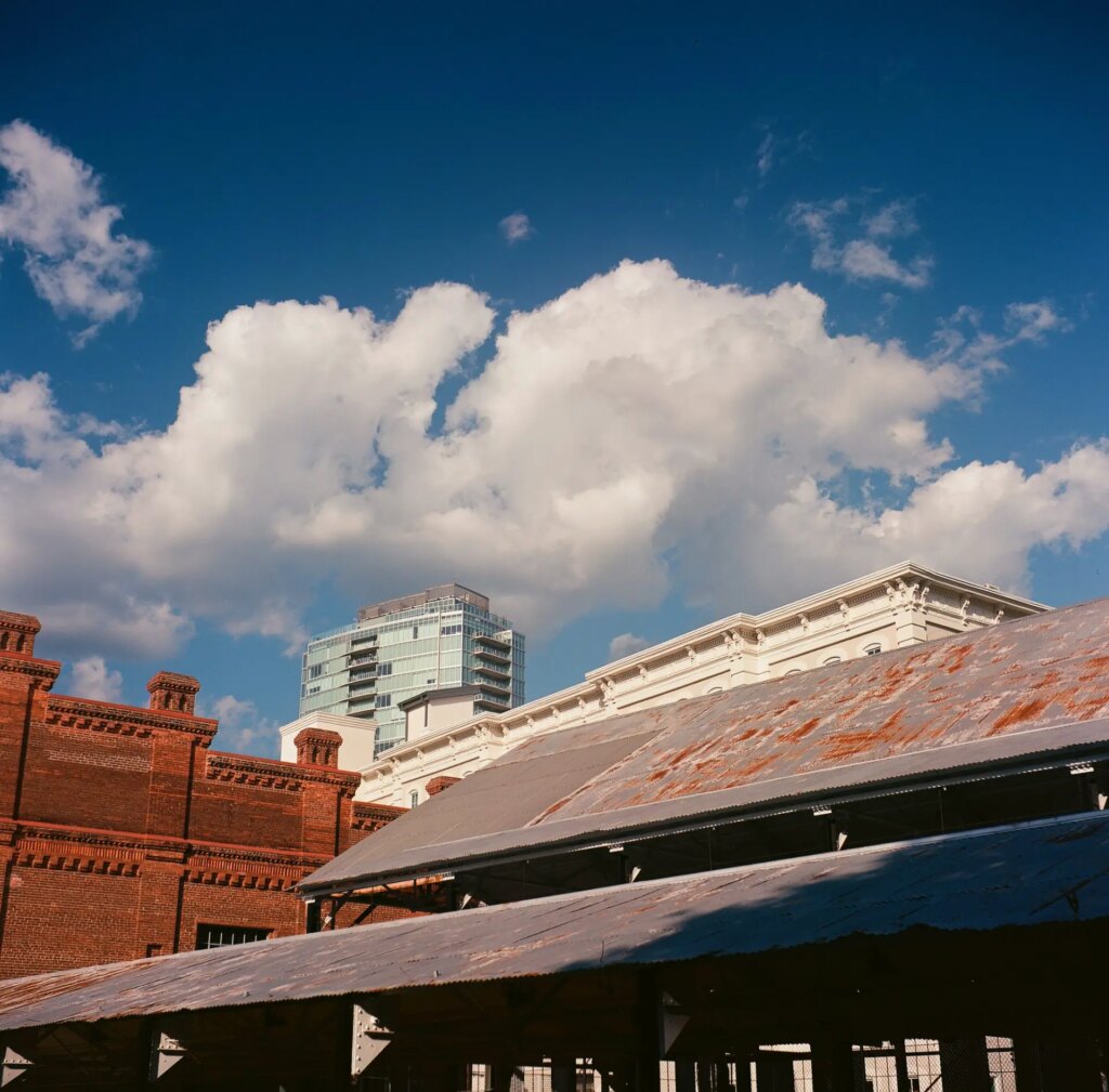 Image shows the tops of four different buildings layered into the background, from a historic brick tobacco warehouse to a modern high-rise apartment building, leading up to a deep blue sky and puffy white clouds.
