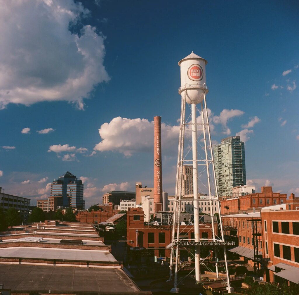 Wide, scenic view of the American Tobacco Campus shot with a Tower Reflex twin lens reflex, from the top level of a parking deck. The old restored Lucky Strike water tower and smoke stack, and roofs of tobacco warehouses, recede into the background of Durham's skyline with a deep blue sky and puffy clouds. 