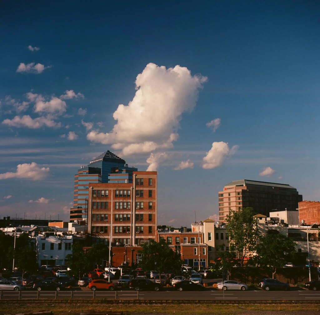 View of the southern edge of downtown Durham, NC, from the American Tobacco Campus north parking deck. The image includes a train track, parked cars, buildings and a deep blue sky with puffy white clouds.