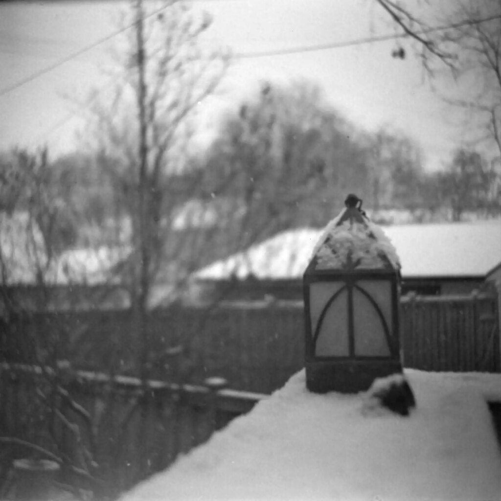 A birdhouse sits on a snow-covered deck behind a house.