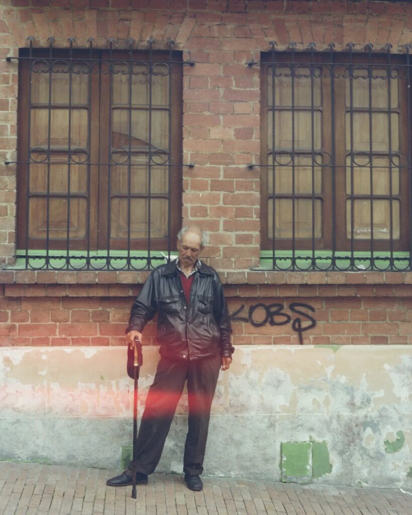 A Bogotano man stands next to a brick building. His eyes are closed and his hips are cocked to one side as he leans on a cane.