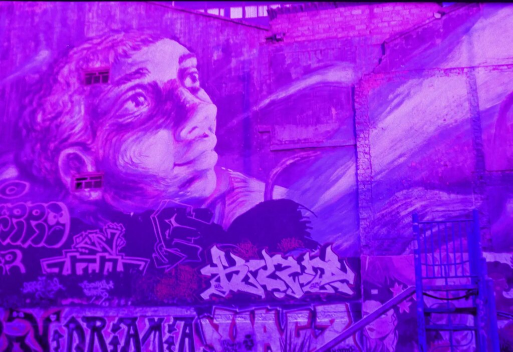 A deep purple photo of a street art mural on a large 3 story building. The mural is of a person looking hop.fully to the sky. Beneath the mural are some other street art throw ups.