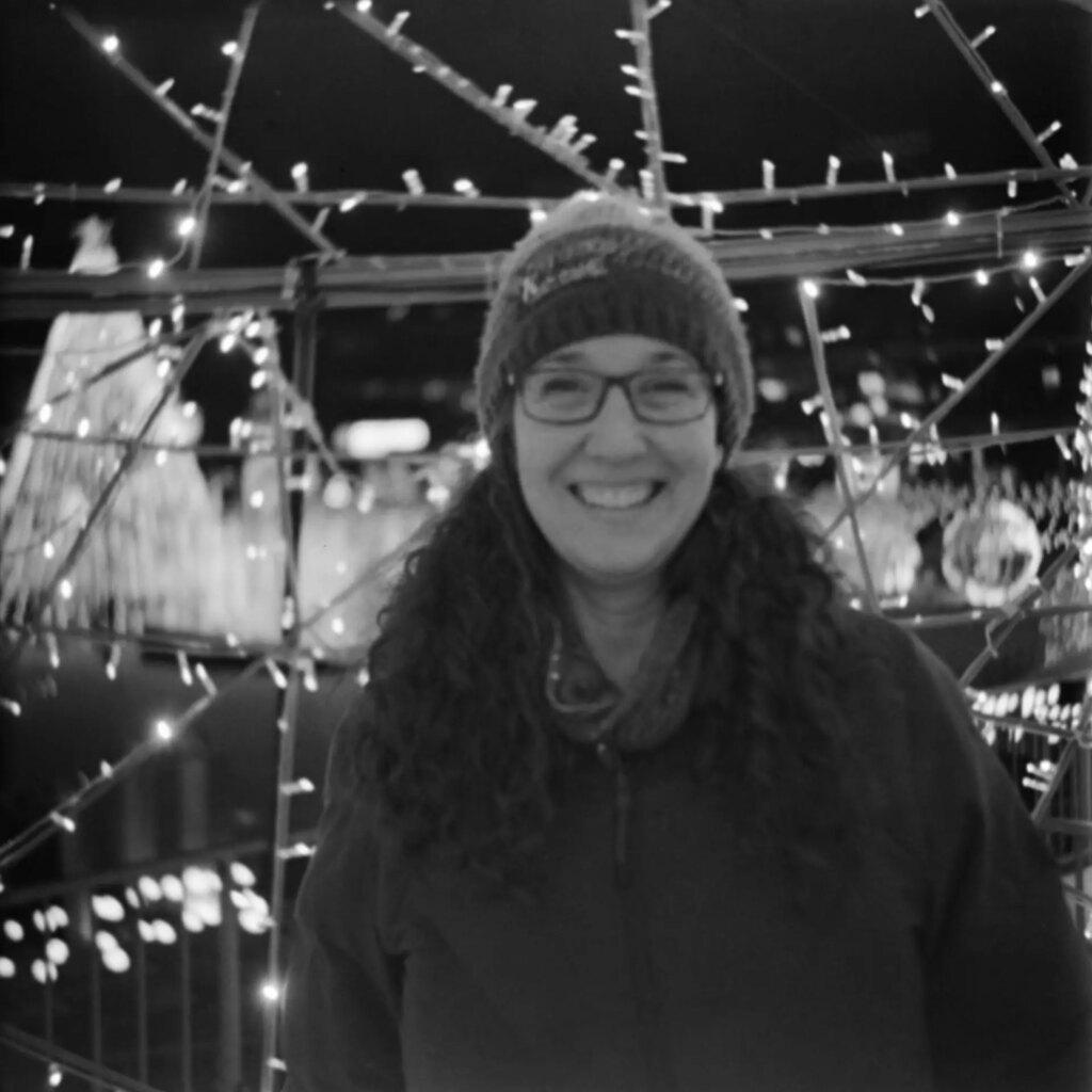 A black-and-white portrait of a woman standing in front of a collection of christmas lights. She is wearing a knit cap and glasses