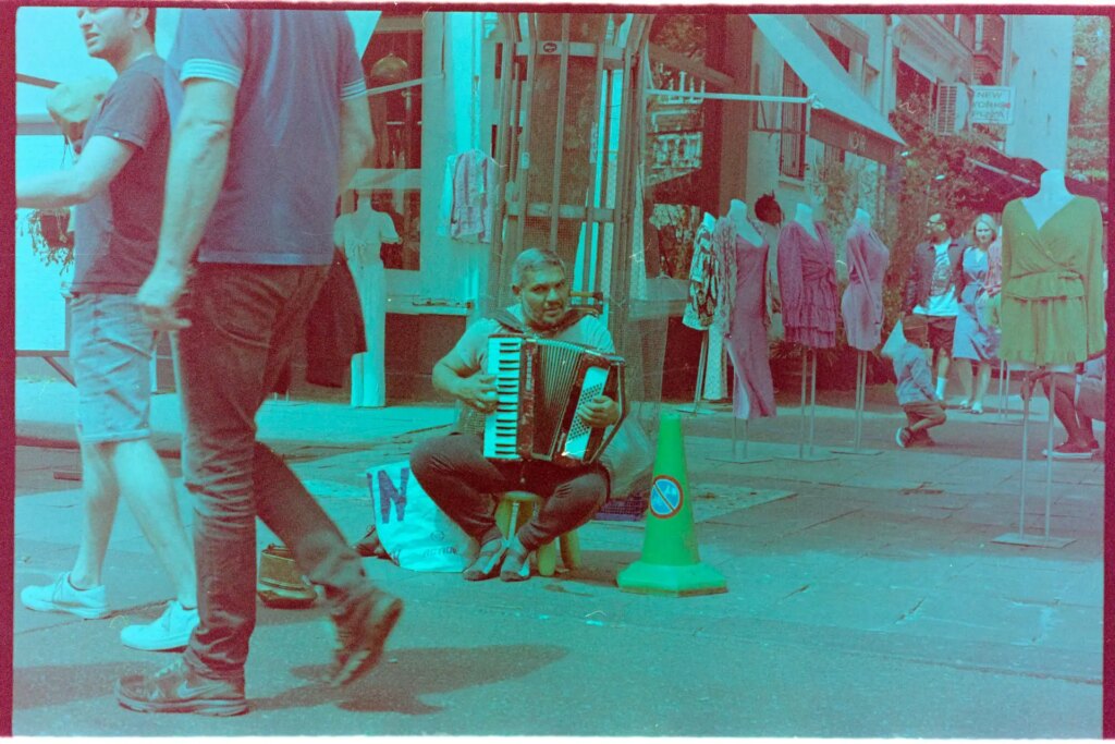 A man sits on the side of the street, and plays accordion for passersby.