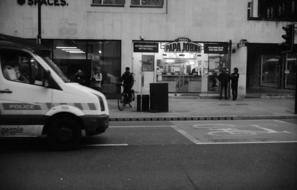 A police van outside a pizza shop, people in the background with obscured faces.