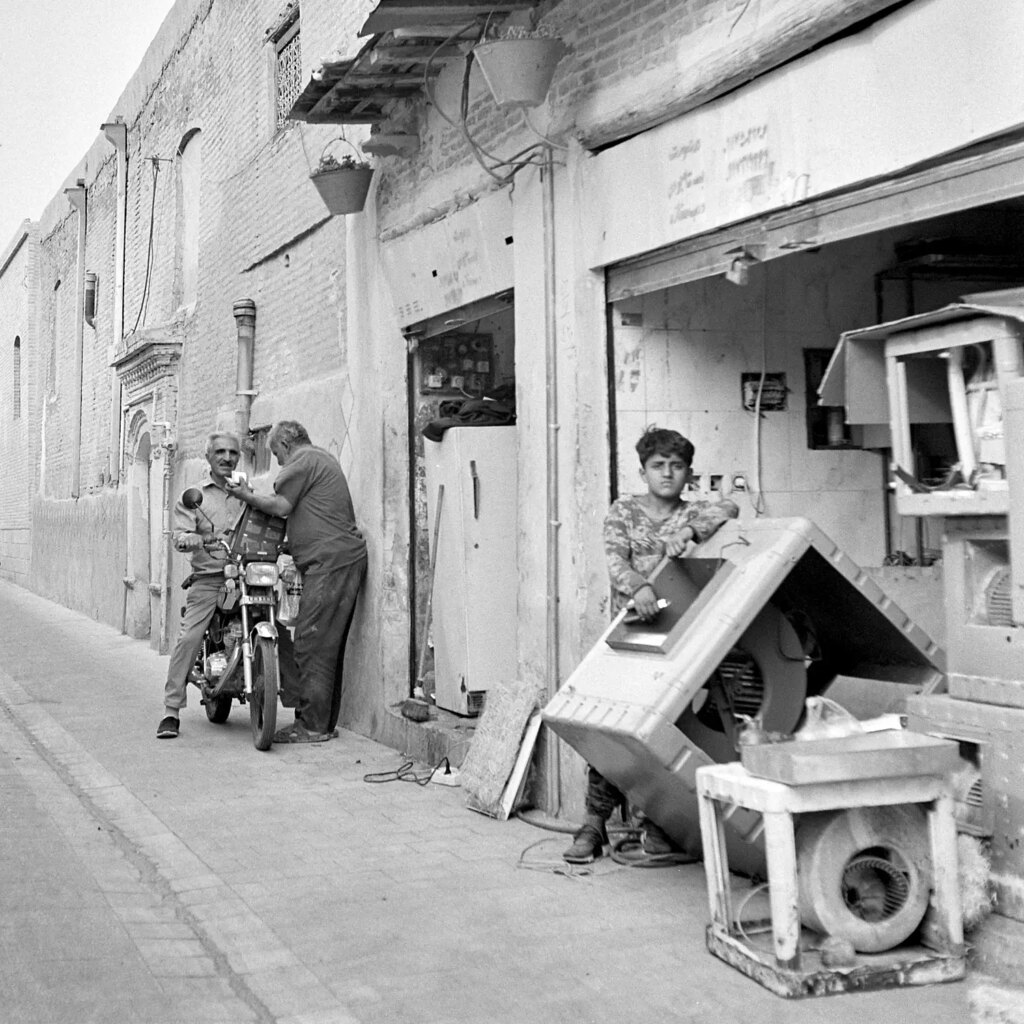 A boy supporting a large appliance that looks like a washing maschine. Few meters behind him a man on a motorcycle talks to another standing next to him.