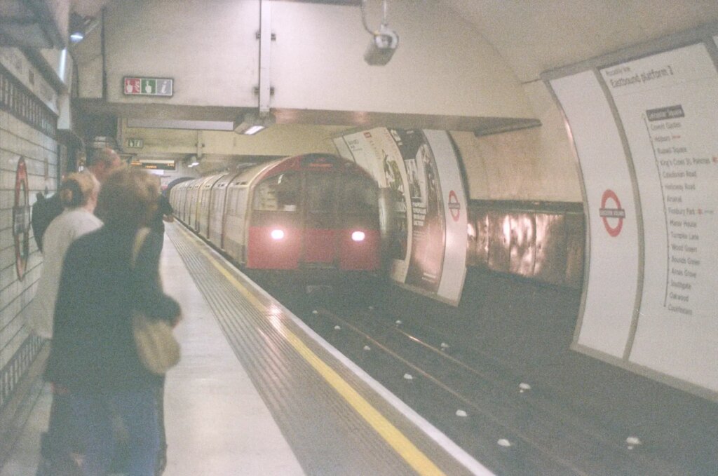 A tube train enters a tube station, with people on the left and signage on the right.