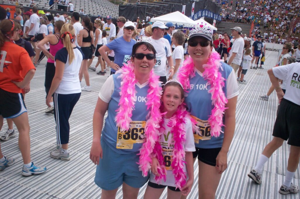 three women in blue and white shirts with tiaras and pink and white feather boas with lots of people in the background in a running finish line studio photo