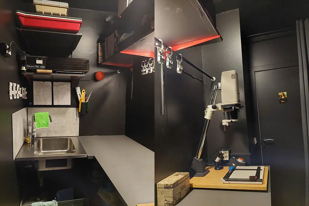 full darkroom, darkroom, enlarger, red light, counter top, sink, trays, print drier, shelves, brackets, lights, black walls, black paint