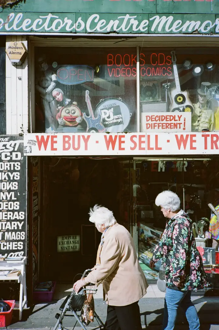 Two Older Ladies pass in front of a vintage shop, with colourful signs advertising and odd trinkets and curios inside