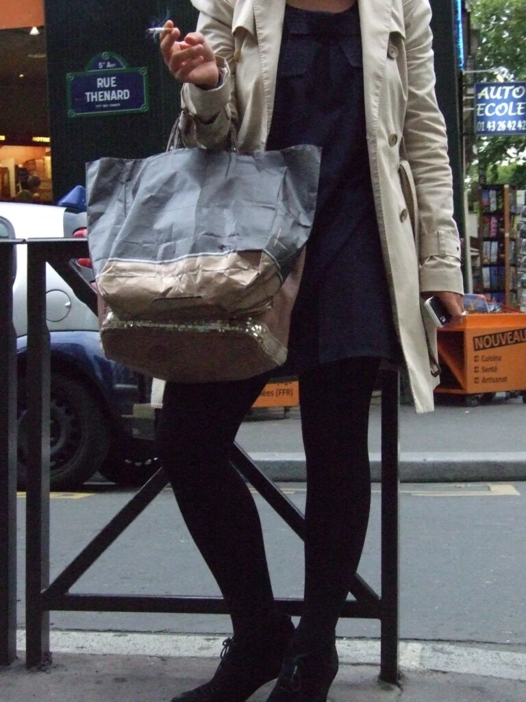 Woman standing on sidewalk, holding bags and lit cigarettete.