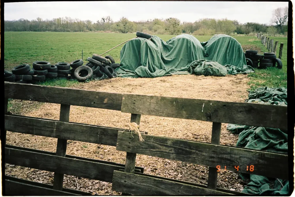 An empty pasture with a fence in the foreground and some straw bales under a tarp as well as some old tires in the distance.