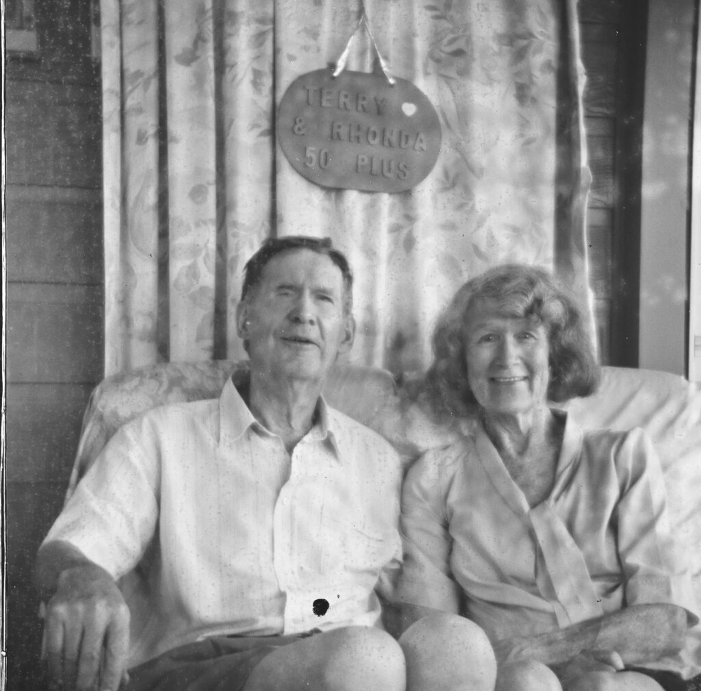 Black and white square image of an elderly couple seated on a couch