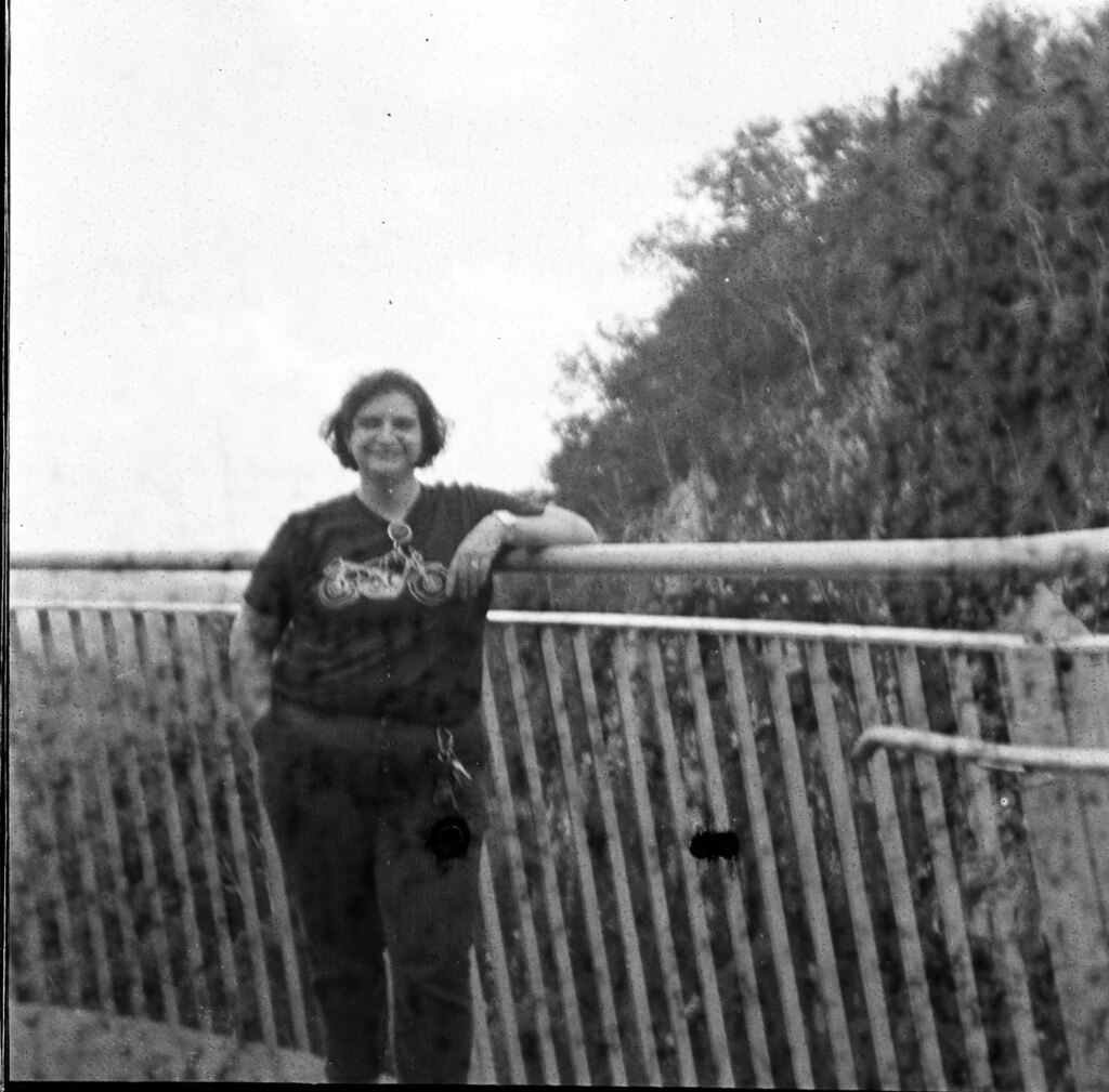 Black and white square image of a woman leaning against a railing. Trees and other foliage visible in the background.