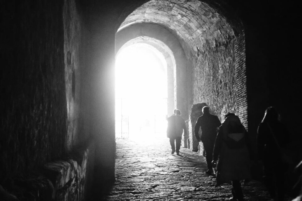 People in a dark passageway walking towards the outside. The contrast of the dark interior and the bright exterior creates an etherial glow.