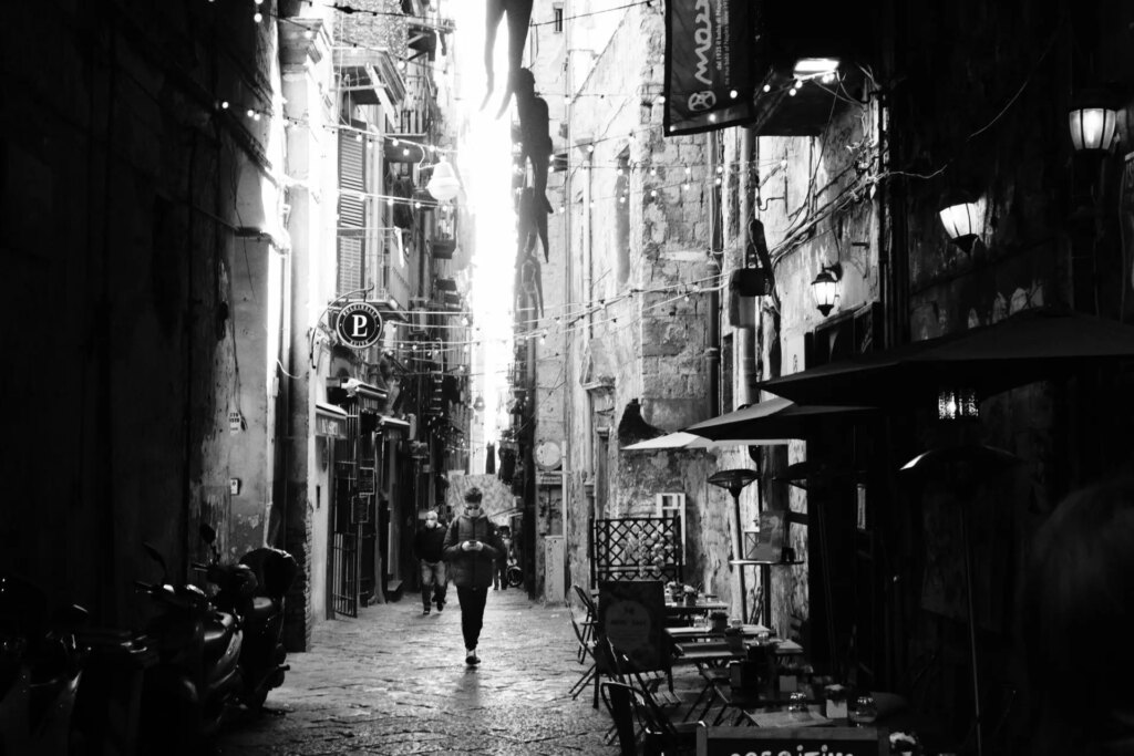 A man stands reading his phone in a dark, very tall, very narrow alley in Napoli. The sun is shining out from somewhere, creating a strong contrast, and lots of glow.