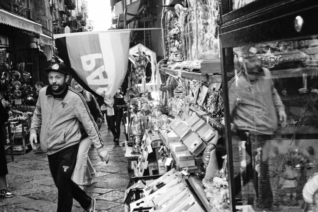 A street full of shops showing their wares. A man walks jauntily under a gay pride flag.