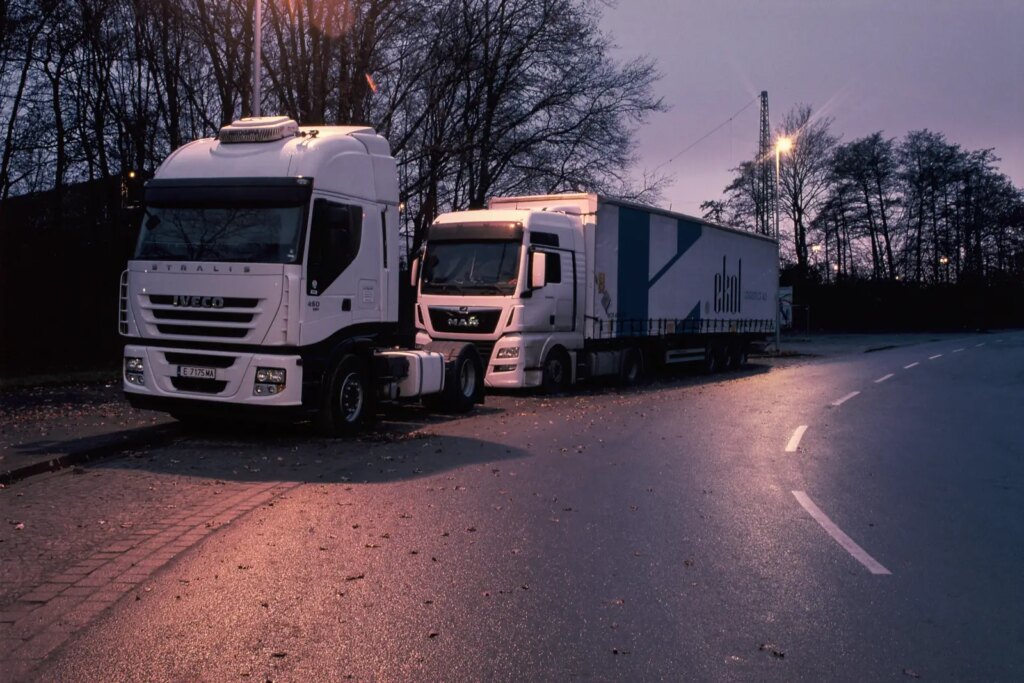 Photograph of two trucks parked at the roadside at dusk. The wet surface of the road reflects the light of the street lamps and shows an orange to purple glow.