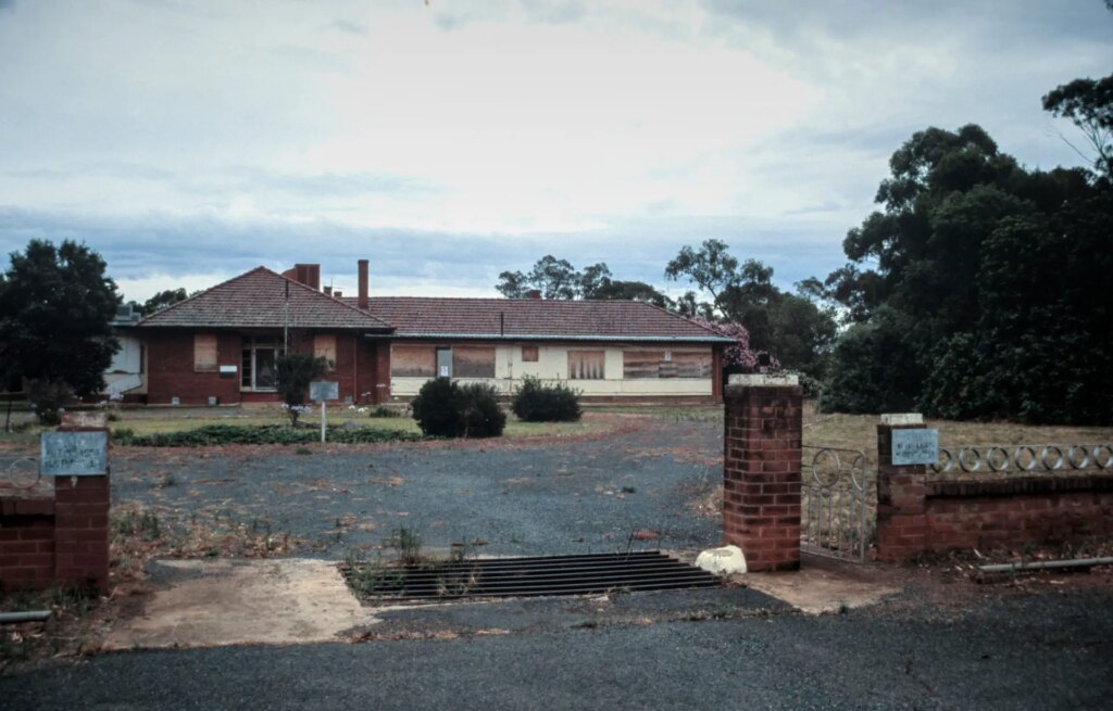 a long single story building with boarded up windows and faded signs on the gate to the large driveway