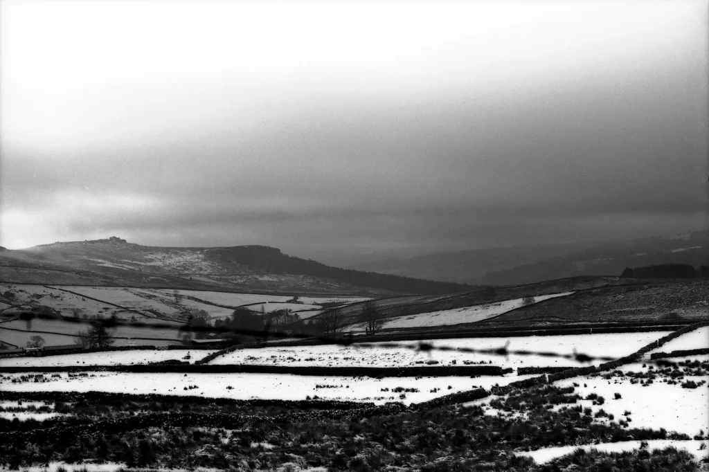 Olympus OM-10 - Features field patches like a patchwork blanket covered in snow with trees and bushes separating each patch. Mountains in the distance can also be seen covered in patches of snow.