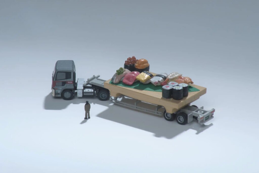 A miniature tractor-trailer pulls a load of oversized pieces of sushi. The diver looks on from beside.