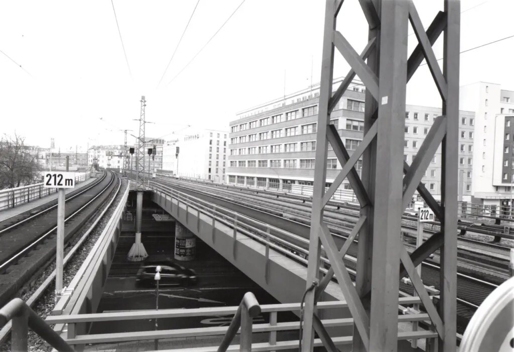 A pylon base is in sharp focus in the fore ground . The power lines traverse the sky above a perfect triangle os lines and barriers. The triaangle forms a viewing window onto the street bellow.