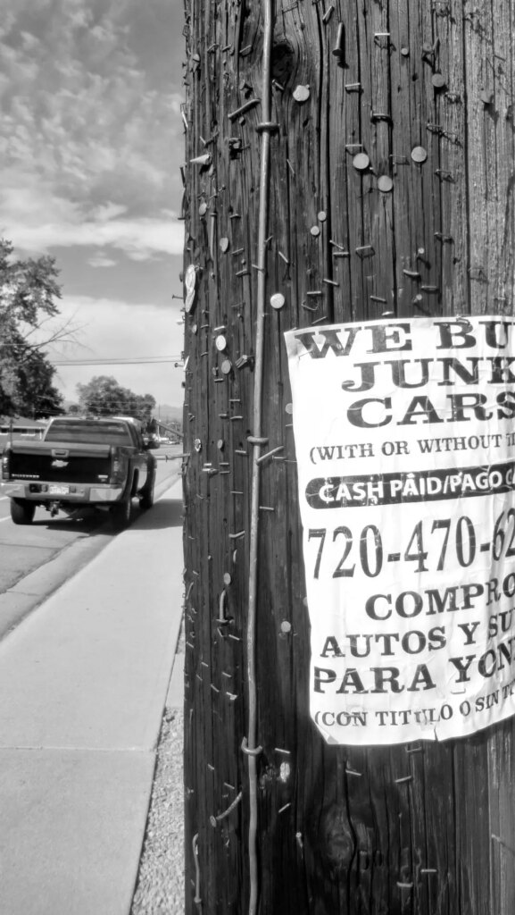 black and white image of an old pickup parked near sidewalk in distance, and near front of frame part of a post with a part of a sign saying "we buy junk cars"