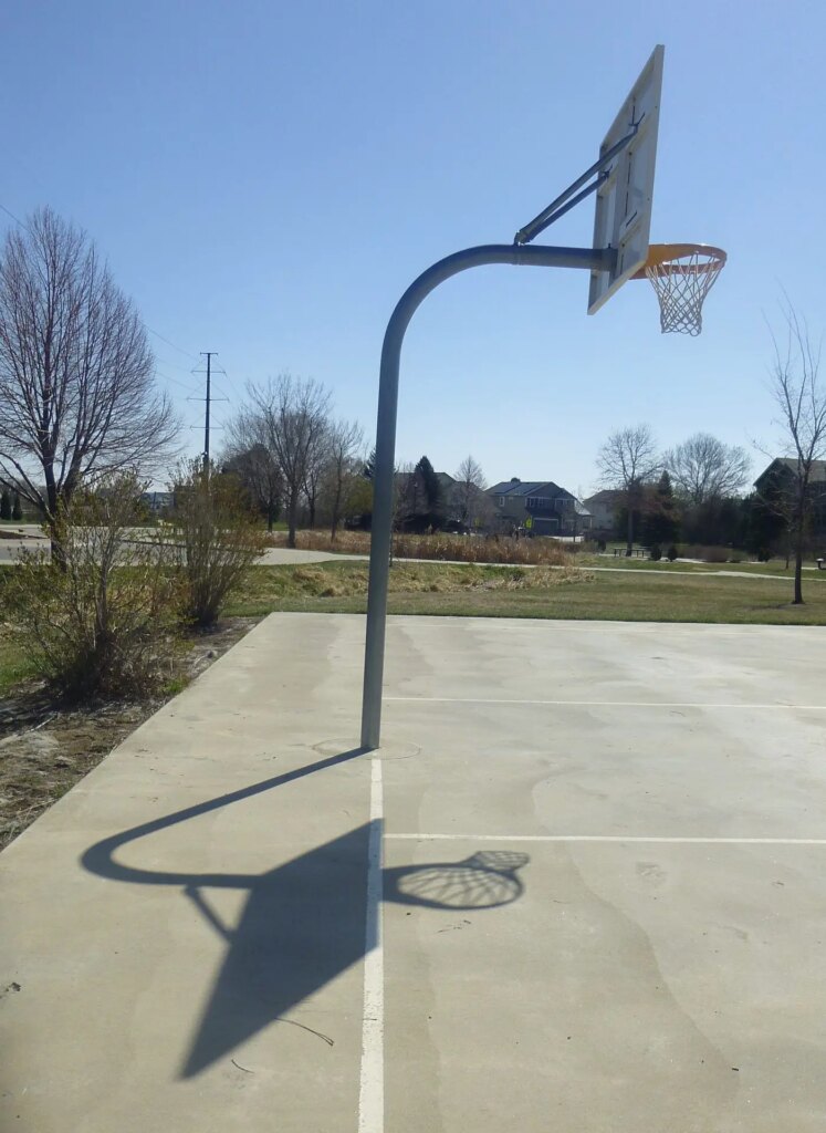 picture of outside basketball goal and background with shadow on a grey concrete playground with winter trees and houses in background, sunny day with blue skey