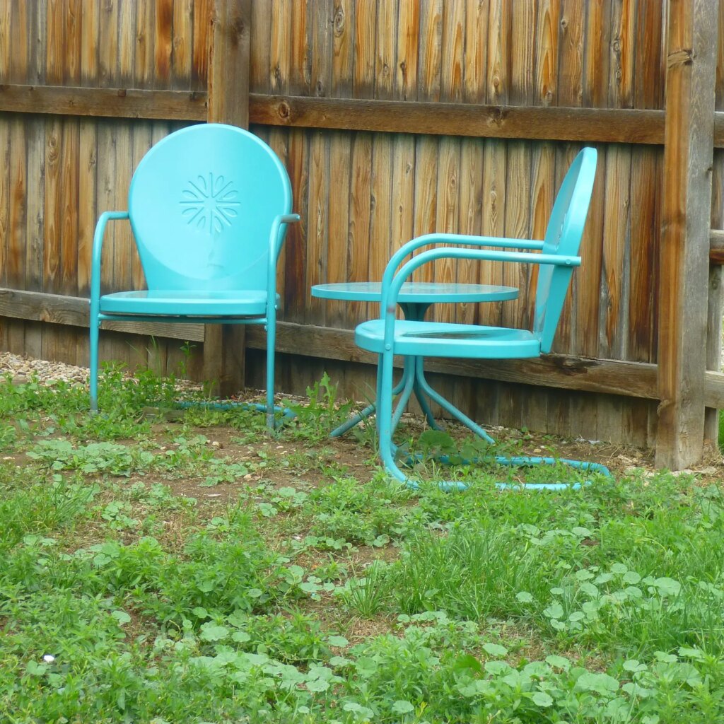 two metal turquoise chairs with grass, dirt, and weeds on ground, and background of wooden fence