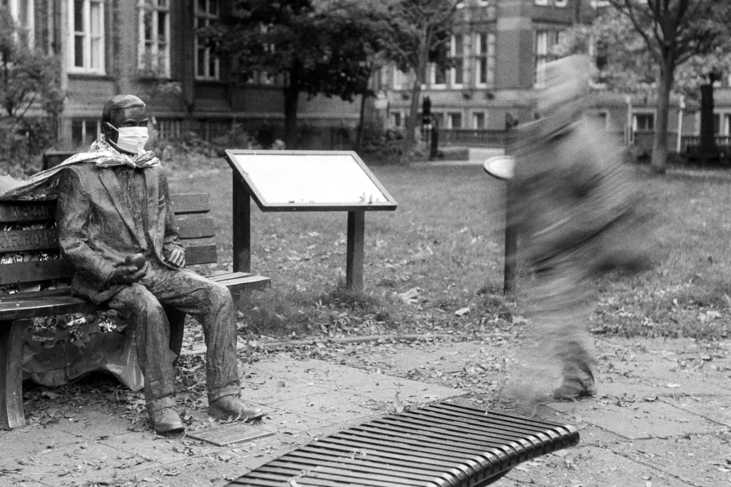 A blurred person walks past a seated statue of Alan Turing that has had a mask put on it