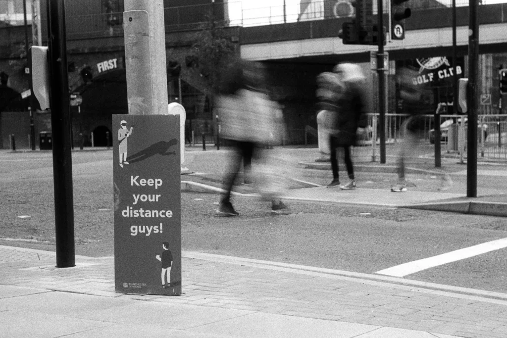 A sign saying 'Keep your distance guys' at traffic lights with blurred people crossing the road