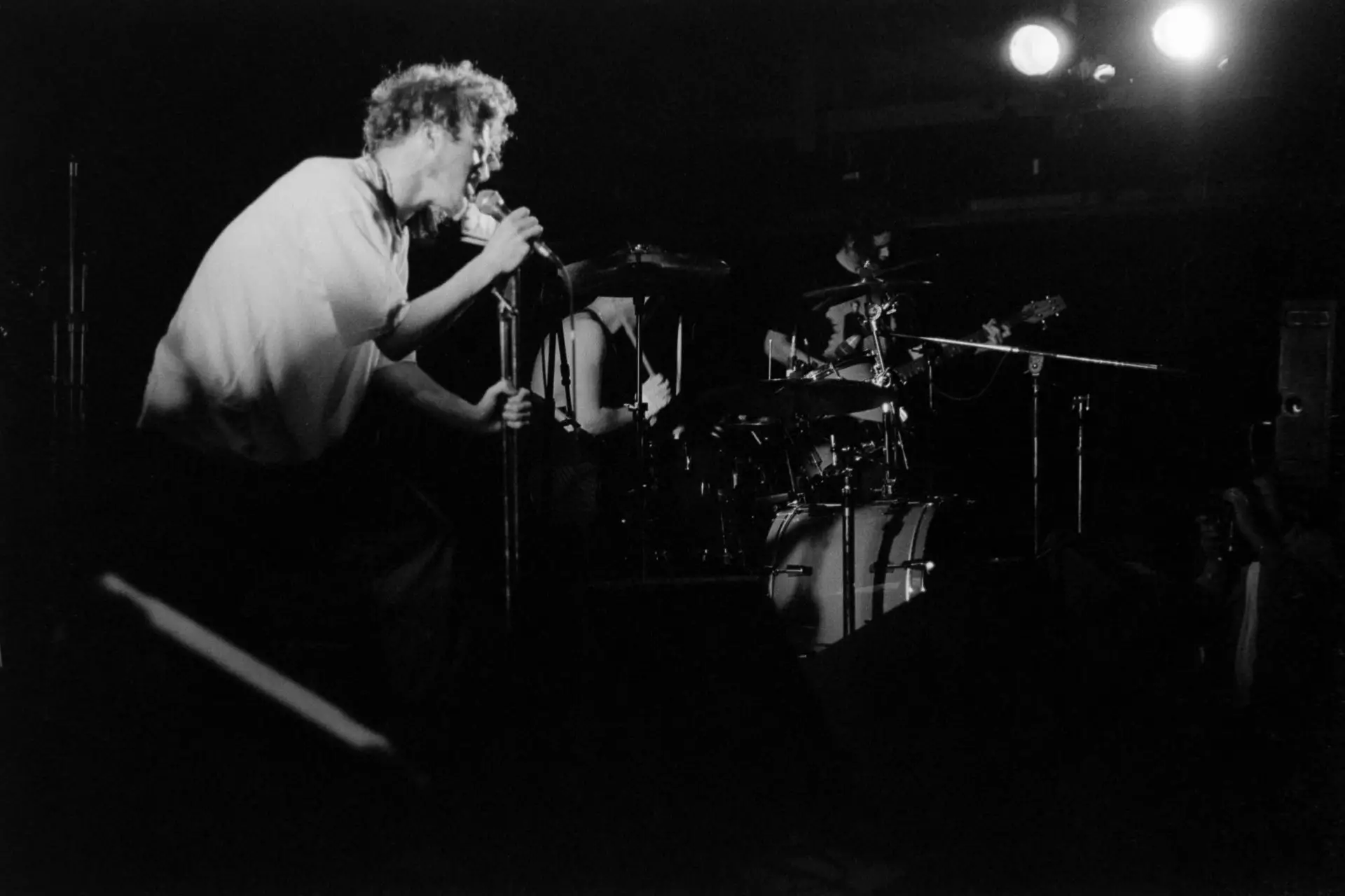 black and white image of a man singing, with a man playing the drums in the background