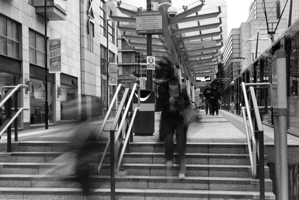 Blurred tram passengers, one of whom is wearing a mask, pass a sign saying wear a face covering. There is a safe distance banner in the background