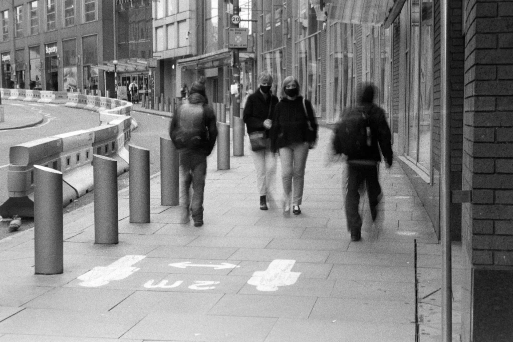 Two masked ladies pass-by two blurred pedestrians. There is a keep two metres apart sign painted onto the pavement