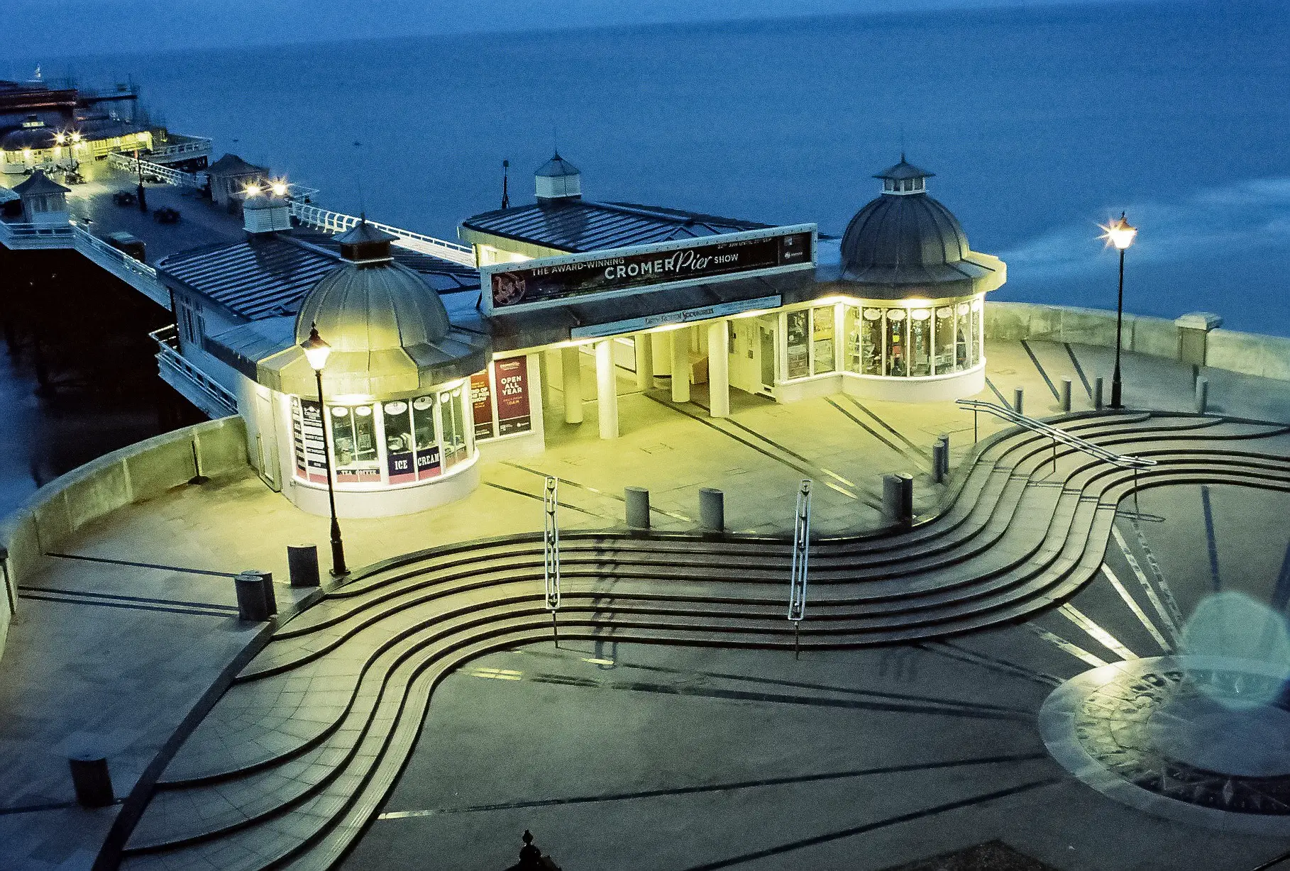 Looking down onto the entrance to Cromer Pier. It's empty of people and the lights tinkle through the darkening night