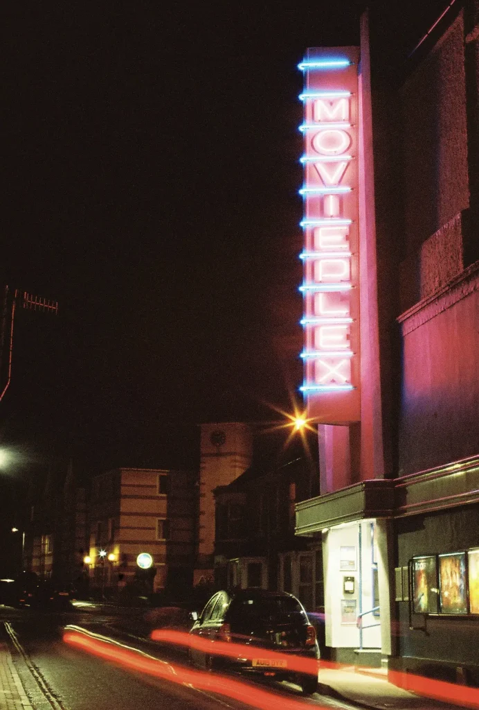 Vertical pink neon sign on the outside wall of a cinema shines in the night. Short red light trail from a moving car underneath.