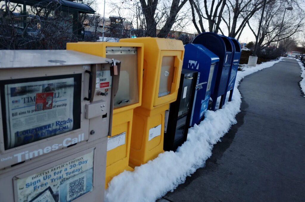 row of newspaper boxes near sidewalk with a bit of snow on ground and trees in background