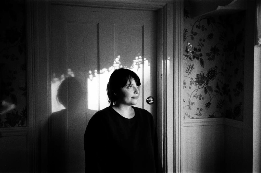 A woman stands in a doorway with curtain shadows around her