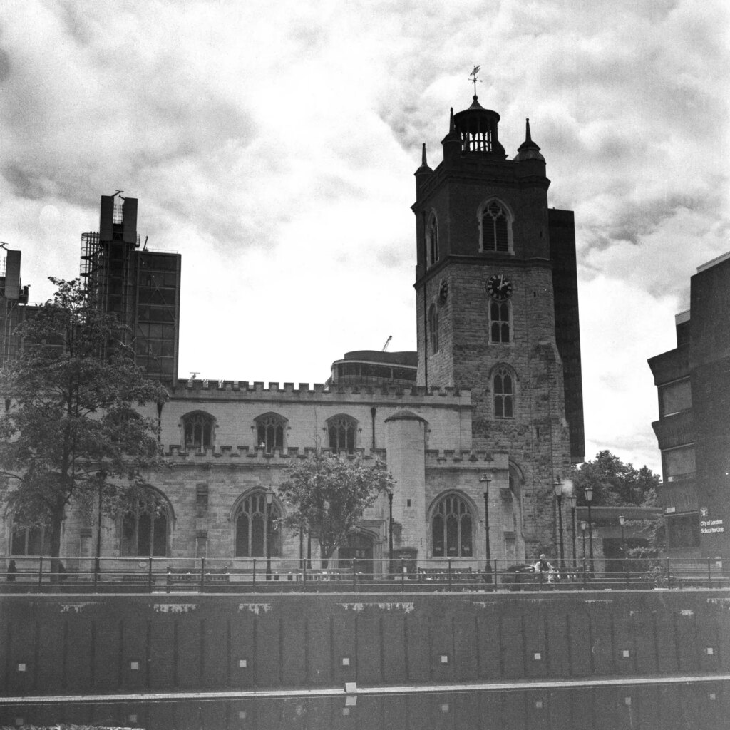 Black and white photo of church in Barbican complex, clouds behind the tower of the church and trees in the foreground