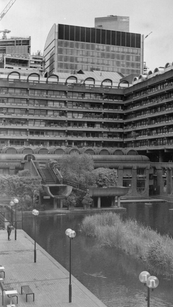 Black and white photograph of the Barbican complex in London. View of residential flats with ornamental lake in the foreground