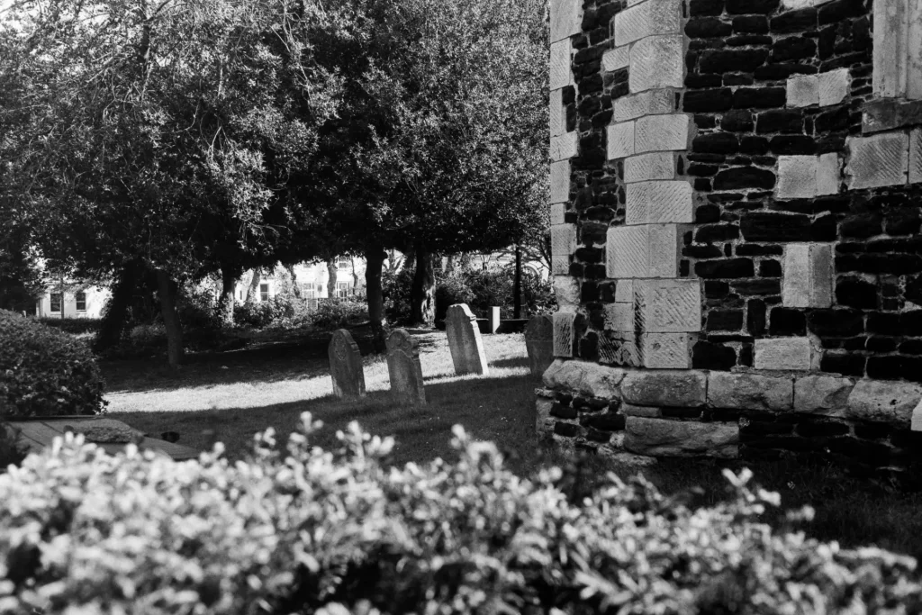 Black and white photograph of gravestones beside a church tower; the light shines on some of the gravestones while others are in shadow. New spring growth in the foreground and the rear