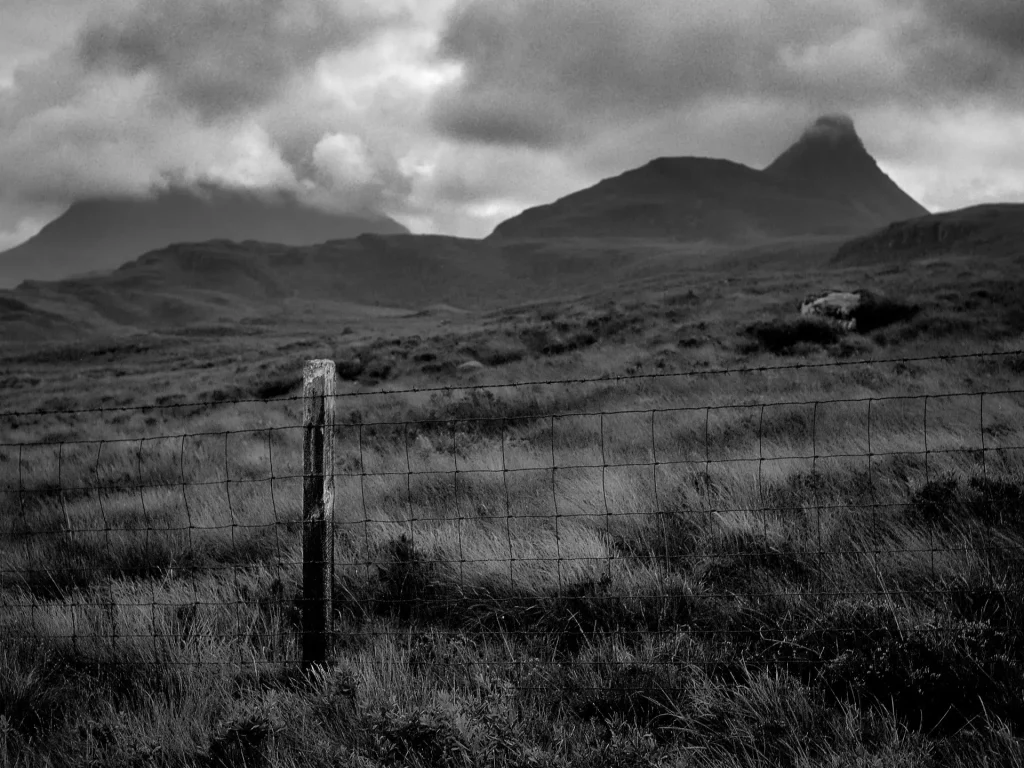 A black-and-white photograph of Stac Pollaidh, a mountain in the Assynt region of Scotland, with a boggy meadow and fence in the foregorund.