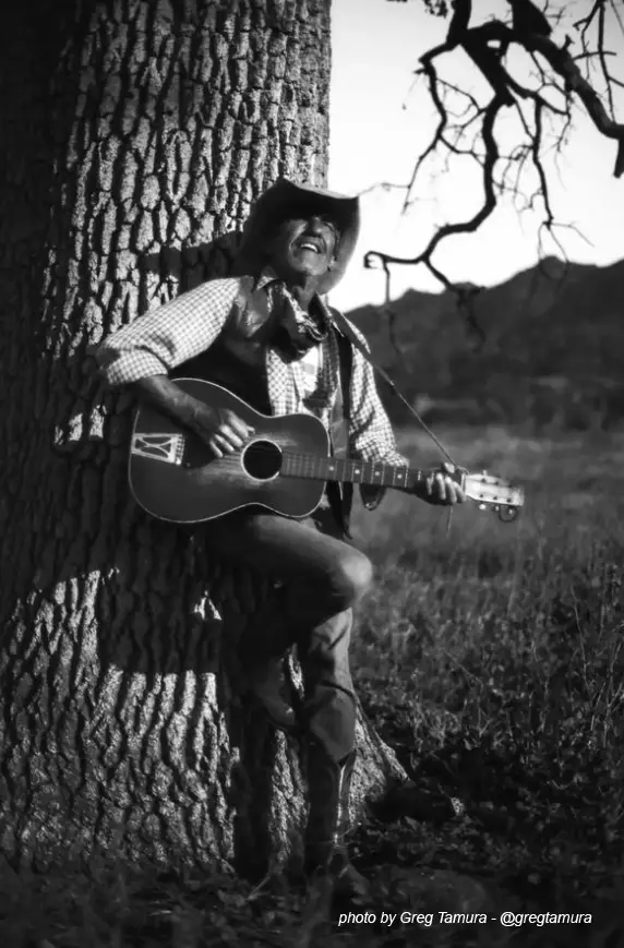 black and white image of man in hat and vest playing guitar leaning against a tree