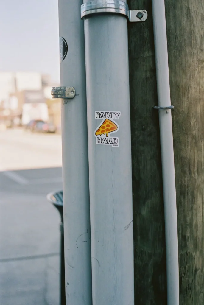 A sticker that reads party hard with a slice of pizza on a pole in the street