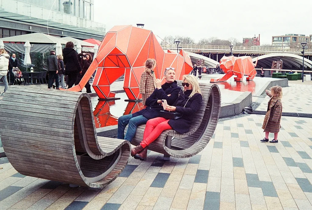 Adult couple sitting on a bench while two young kids play around Battersea Power Station park features